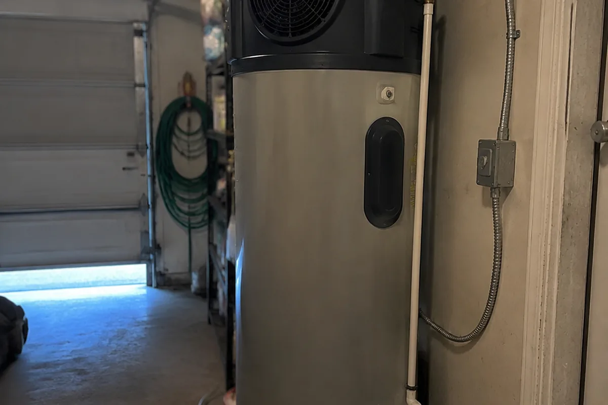 A residential heat pump water heater installed in a garage with visible electrical conduit, condensate line, and duct connections