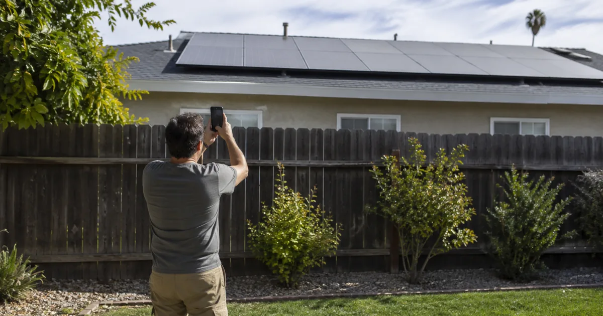 A city building inspector on a laptop conducting a video call inspection while a homeowner in the background points a phone camera toward a rooftop solar array