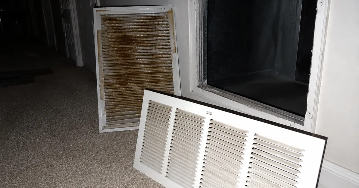A furnace return air grille removed and set on the carpet, its pleated filter stained with a sticky brown film, the clean wall opening visible behind it