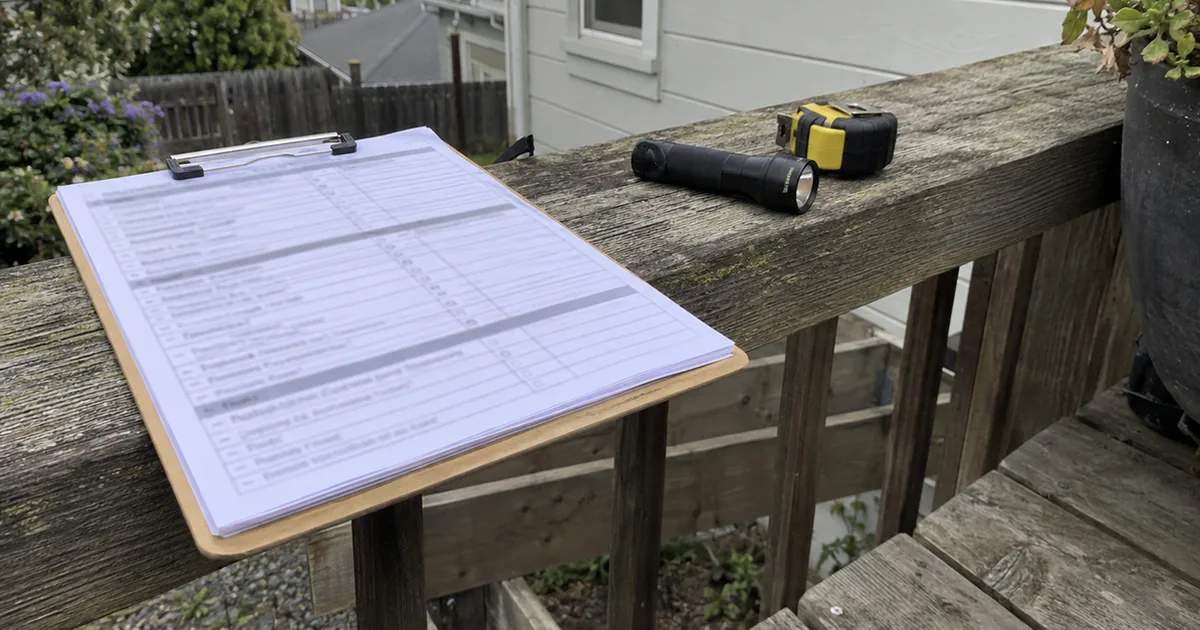 A clipboard with an open inspection checklist resting on a weathered wooden deck railing next to a flashlight and a tape measure, overcast daylight