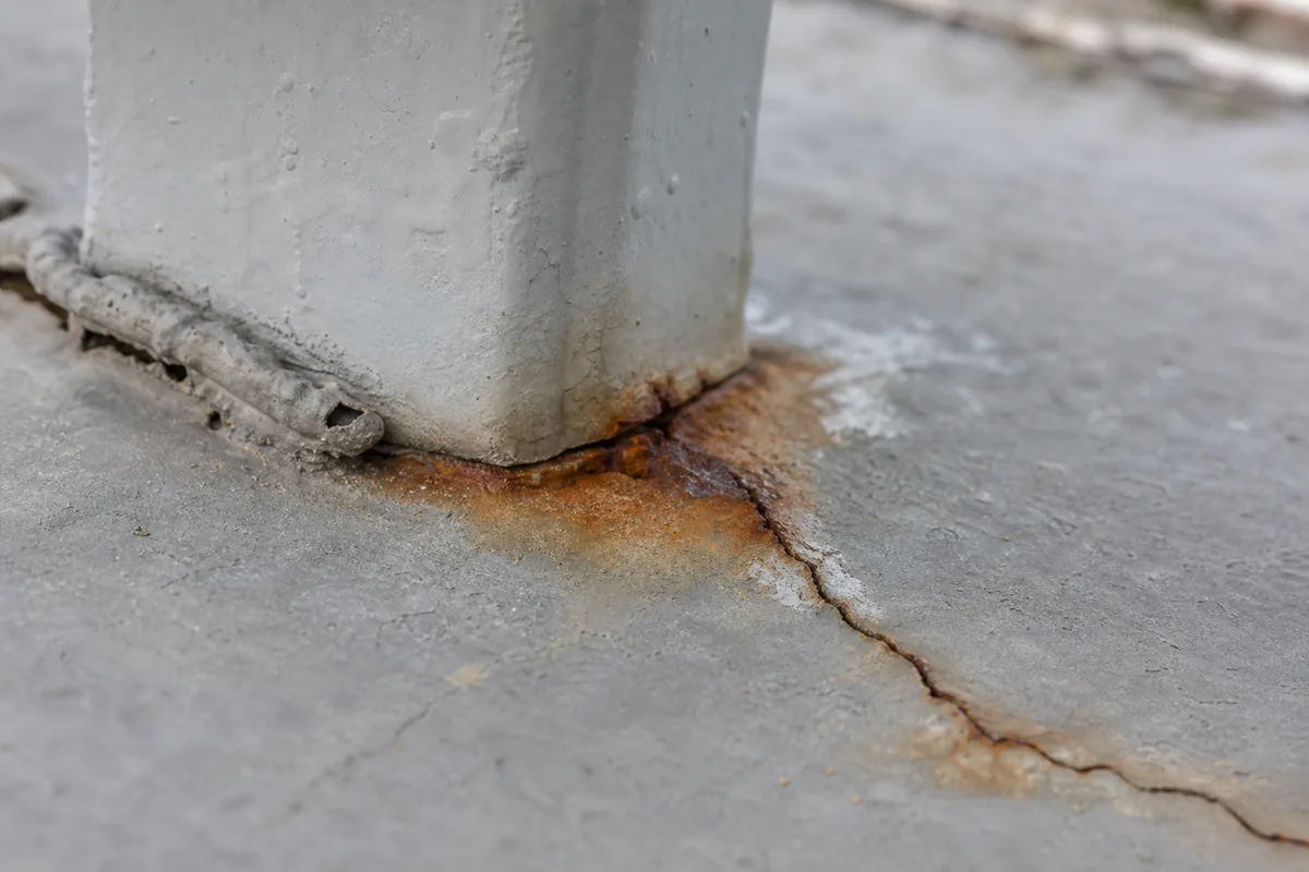 Close-up of a rusted metal balcony railing post where it penetrates cracked elastomeric deck coating with orange-brown rust bleeding into the surface