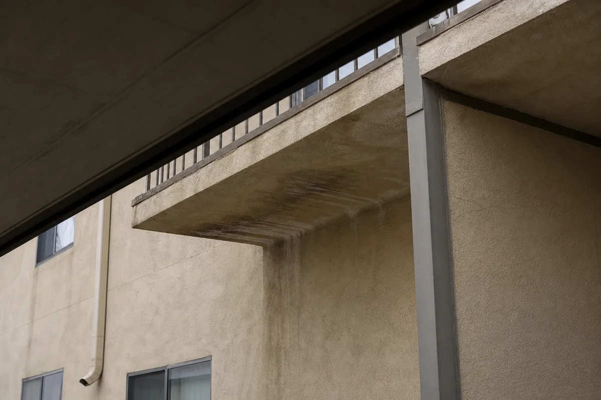 Dark water stain and efflorescence streaking down the beige soffit beneath an elevated walkway on an older stucco multifamily building
