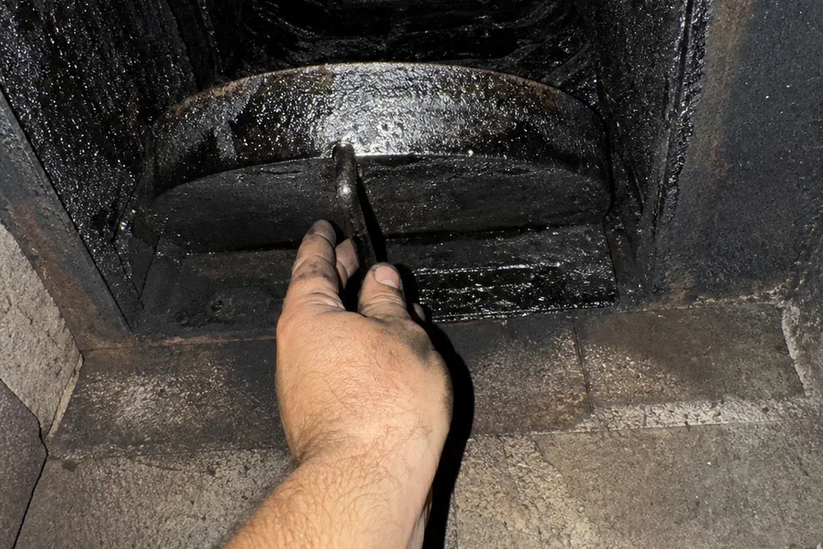 Hand reaching up into a sooted fireplace throat to push the metal damper lever open, glazed creosote coating the masonry