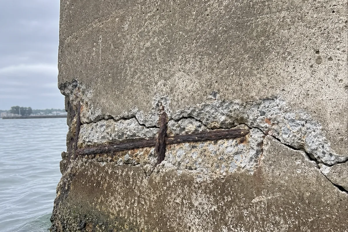 Close-up of a concrete seawall face at the waterline with spalling concrete and rust-stained exposed rebar