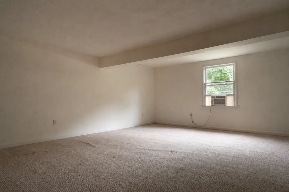 Interior of a converted garage used as a room, with a low flat ceiling, a painted-over former garage-door header beam, a slightly lower slab floor under thin carpet, and a window AC unit