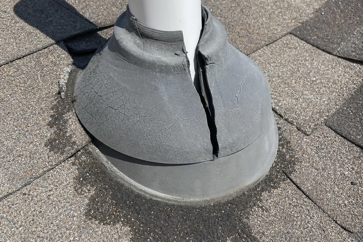 Close-up of a cracked, dried rubber plumbing vent boot on an asphalt shingle roof with staining below it