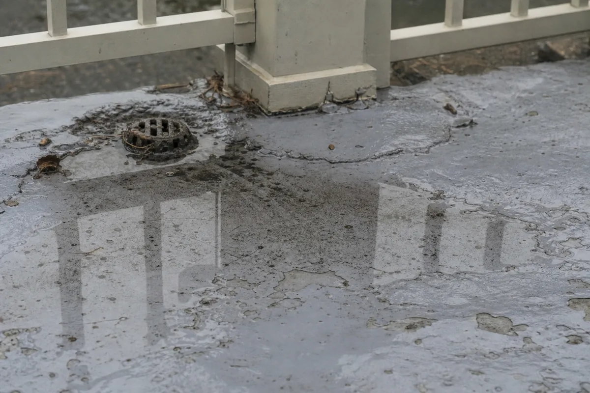 An elevated deck surface where the waterproof coating has cracked and blistered, with standing water pooled against a clogged drain and the base of a railing post