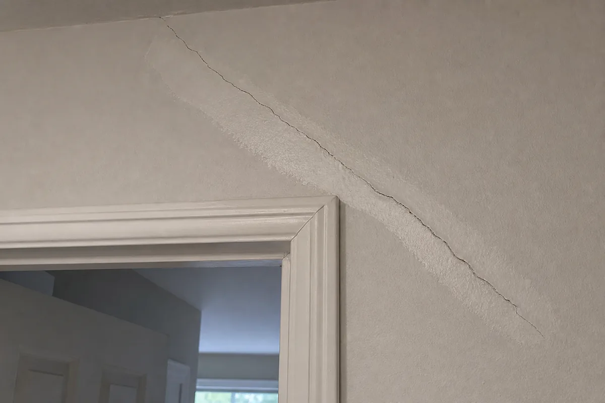 A wide diagonal crack running from the upper corner of an interior doorway across painted drywall, filled and repainted once and reopened along the same line, beside an out-of-square door frame