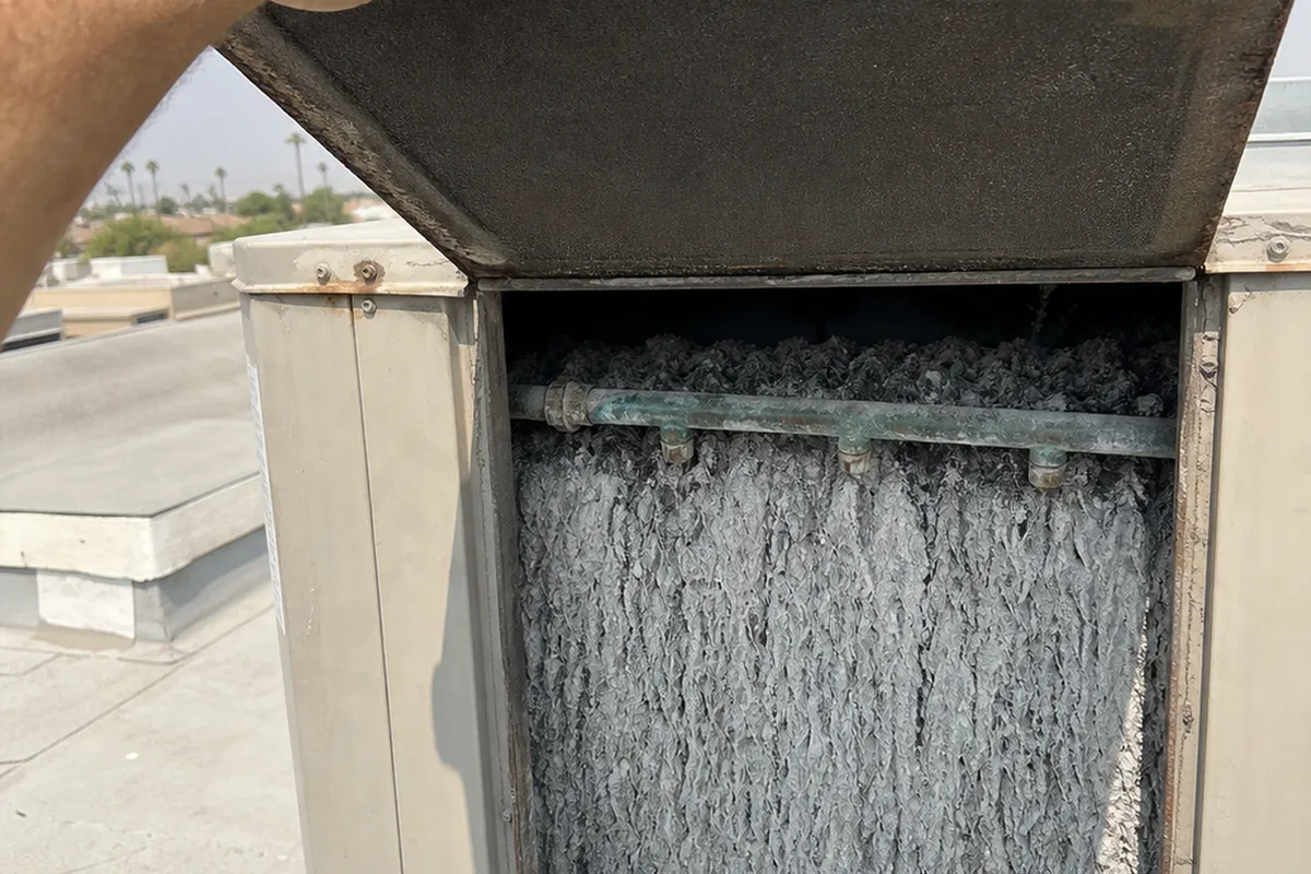 A technician’s hand lifting the access panel on an evaporative cooler cabinet to reveal a mineral-scaled cooling pad and a corroded water distribution line inside