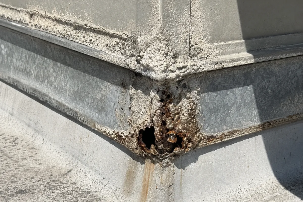 Extreme macro of a rooftop evaporative cooler cabinet’s lower seam with thick mineral scale and rusted-through pinhole corrosion at the corner joint
