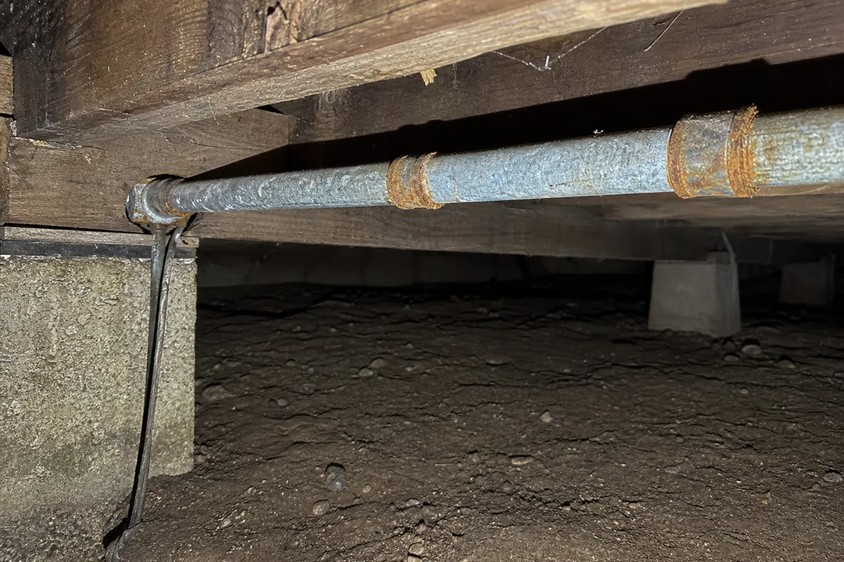 Galvanized steel supply pipe with rust blooming at the threaded fittings, running along the joists of an old crawlspace