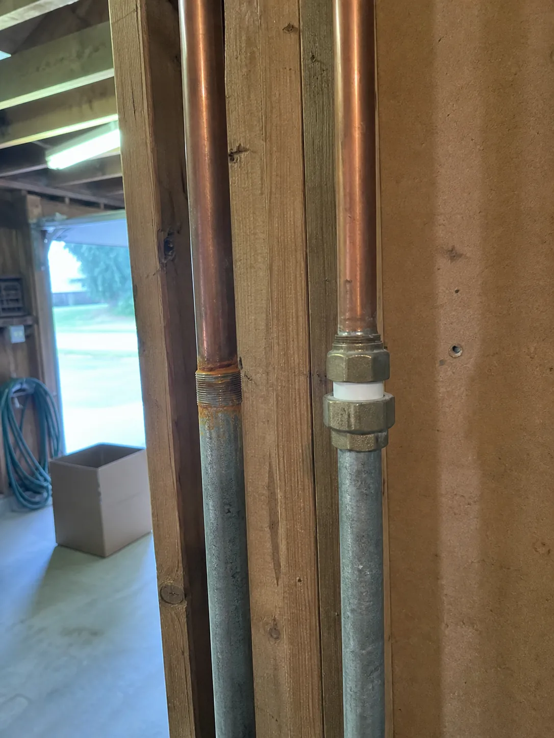 Two adjacent pipe transitions on a garage wall, one a bare threaded copper to galvanized joint with rust staining and one fitted with a proper dielectric union, in dim mixed garage light
