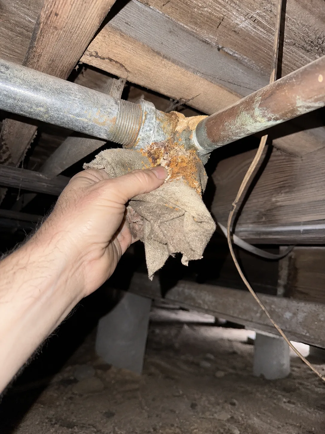 Shot from below looking up beneath a raised beach cottage, a bare hand wiping loose corrosion crust off a copper to galvanized threaded junction with a rag, hard on camera flash and deep black falloff behind the floor framing