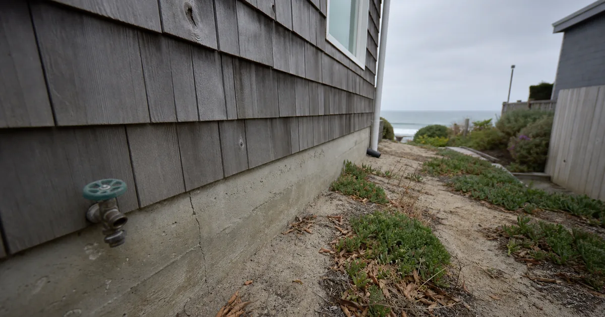Weathered wood-shingle coastal cottage foundation on sandy ground with the ocean visible in the background