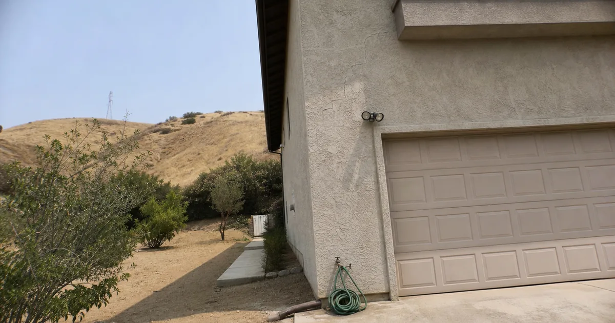 Stair-step stucco crack above a garage on an inland hillside tract home with dry hills behind it