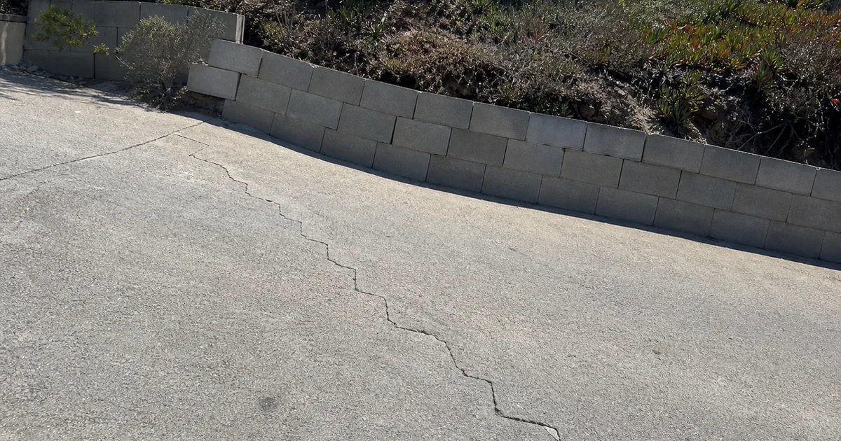 Diagonal stair-step crack across a hillside driveway with a leaning concrete block retaining wall behind it