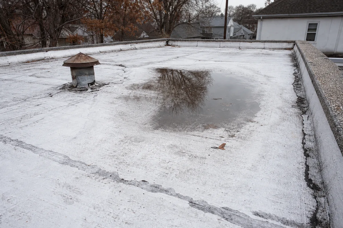 A flat low-slope residential roof with a worn whitish coating, shallow ponded water in a low spot, and cracking, patched seams near a gravel-stopped parapet