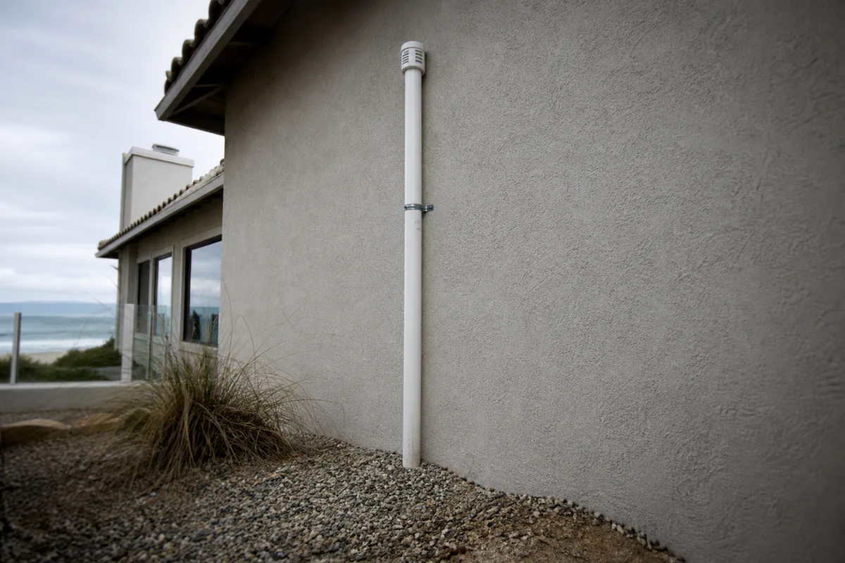 White PVC soil-gas vent riser on a coastal stucco wall with the ocean visible in the background
