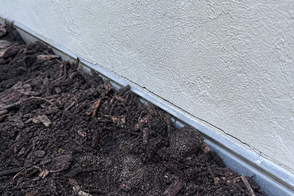 Base of a new stucco wall where the metal weep screed is partly buried in fresh soil and bark mulch, with a hairline crack starting at the screed line