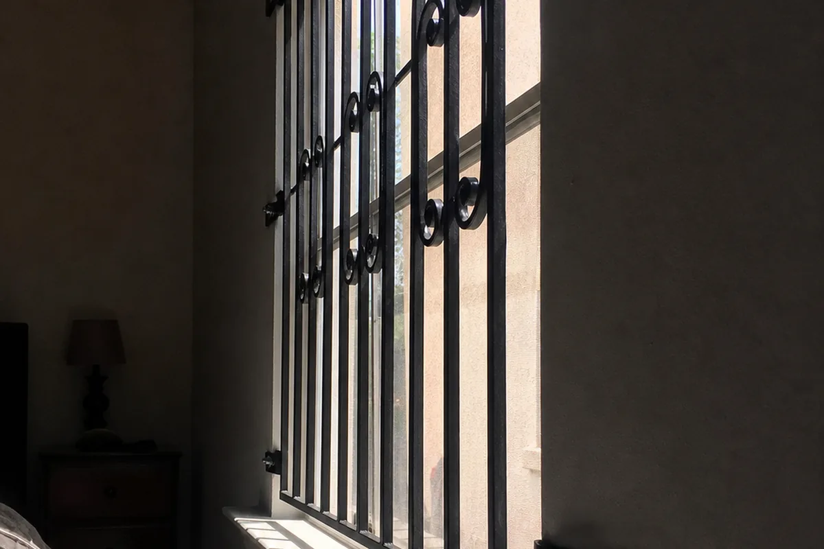 Fixed wrought-iron security grille bolted over a bedroom window with no interior release, hard bar-shadows across the sill