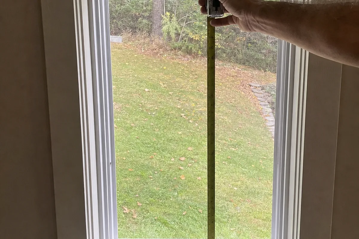 Hand holding a tape measure stretched across the clear opening of a fully opened bedroom window in daylight