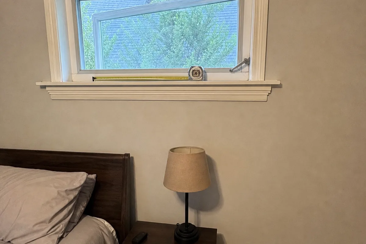 Casement bedroom window cranked open only partway with a high sill, a tape measure set down across the small clear opening