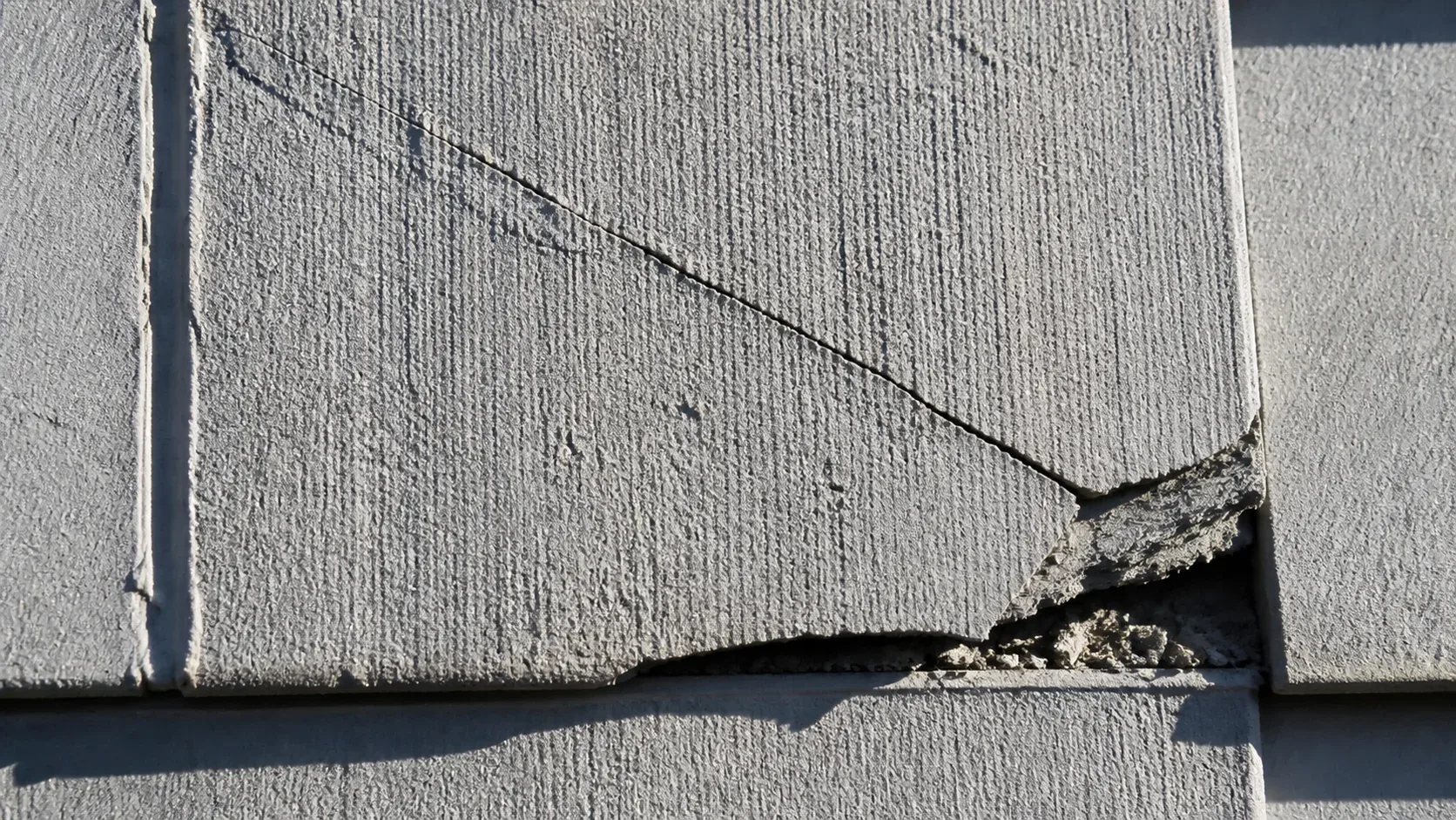 Extreme close-up of a cracked and spalling gray asbestos cement shingle panel on a Panorama City exterior wall, with a small missing corner and fragments below the crack
