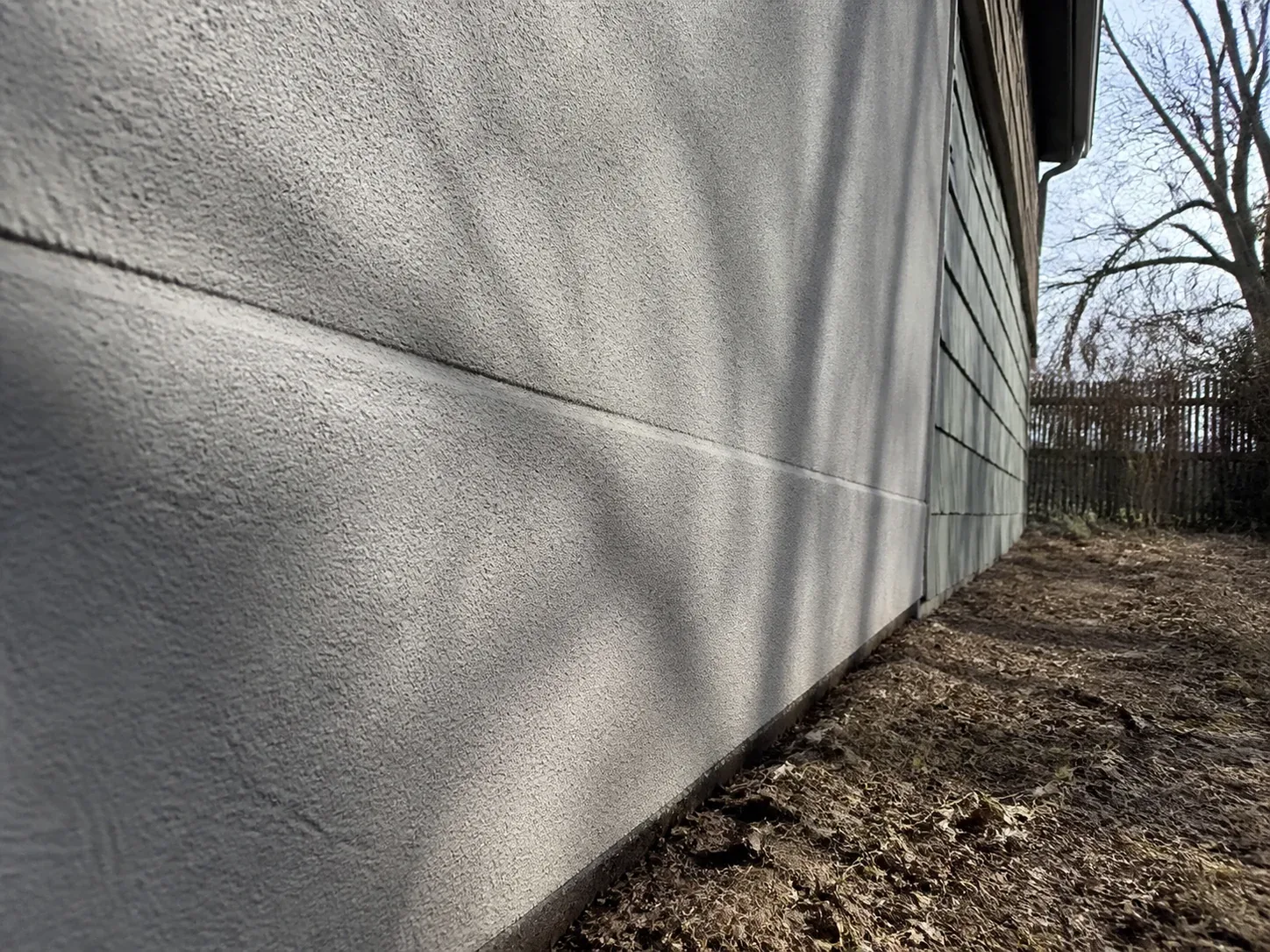 Camera sighting along an exterior wall at a raking angle where smooth stucco transitions into older gray asbestos cement shingle siding at a rear corner of a Panorama City home