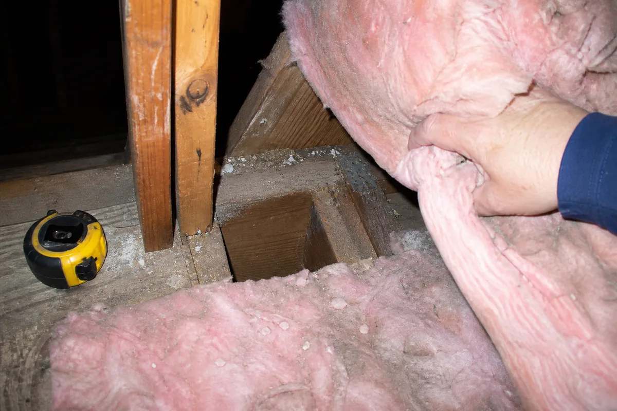 A hand pulling back attic insulation to expose an open, unblocked stud bay at the top plate near an addition’s tie-in point, tape measure set down in frame