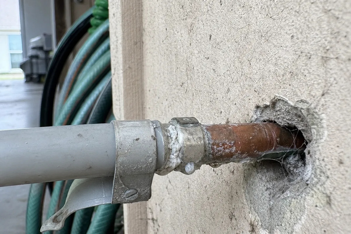 Gray polybutylene supply pipe crimped to a copper stub at a garage main water entry, mineral crust weeping at the band