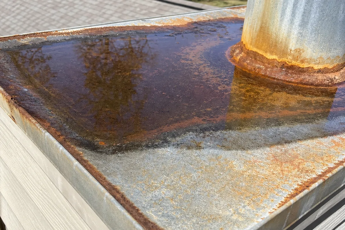 Rusted sheet-metal chase-top pan with standing water pooled on the flat surface, no slope or cricket