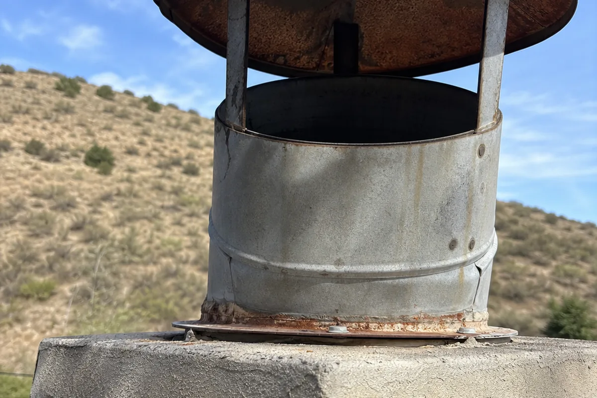 Rusted rain cap on a round metal chimney flue with the spark-arrestor mesh screen missing, exposing the open flue
