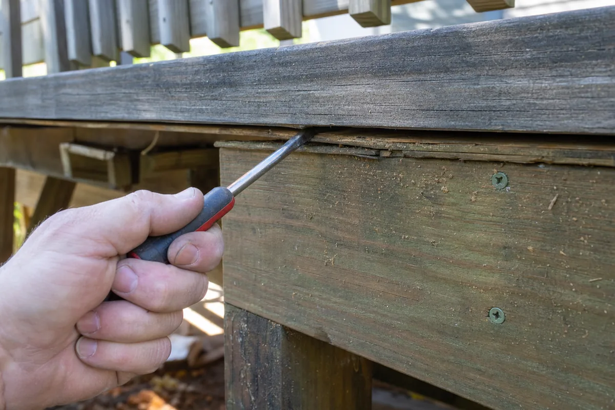 A hand pressing a pry tool into the gap between a deck ledger board and the rim joist to test for movement