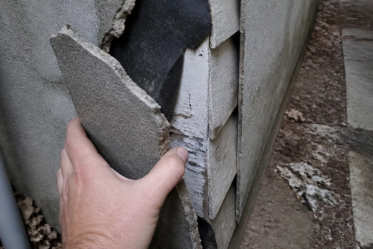 A gloved hand lifting a loose drummy section of stucco away from the wall at a corner, revealing original wood siding and building paper behind it