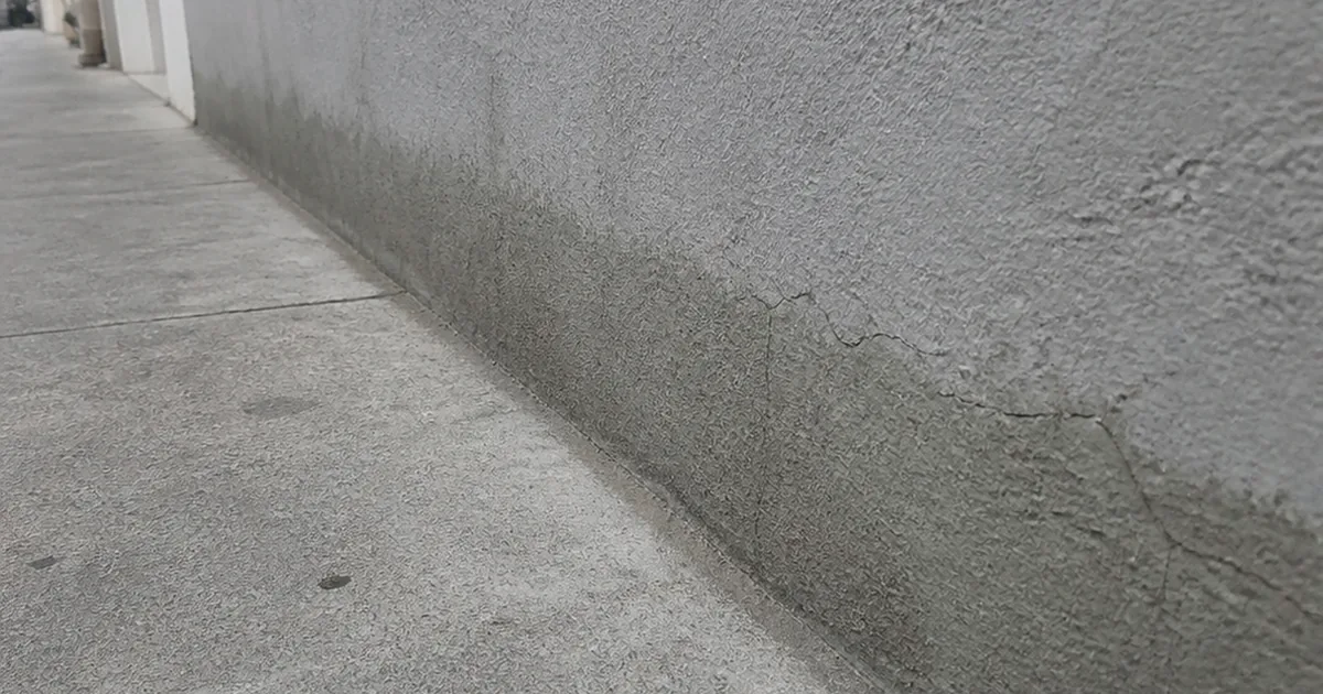 Stucco running straight down into the sidewalk at the base of a San Francisco row house with hairline cracking and a damp tidemark along the bottom
