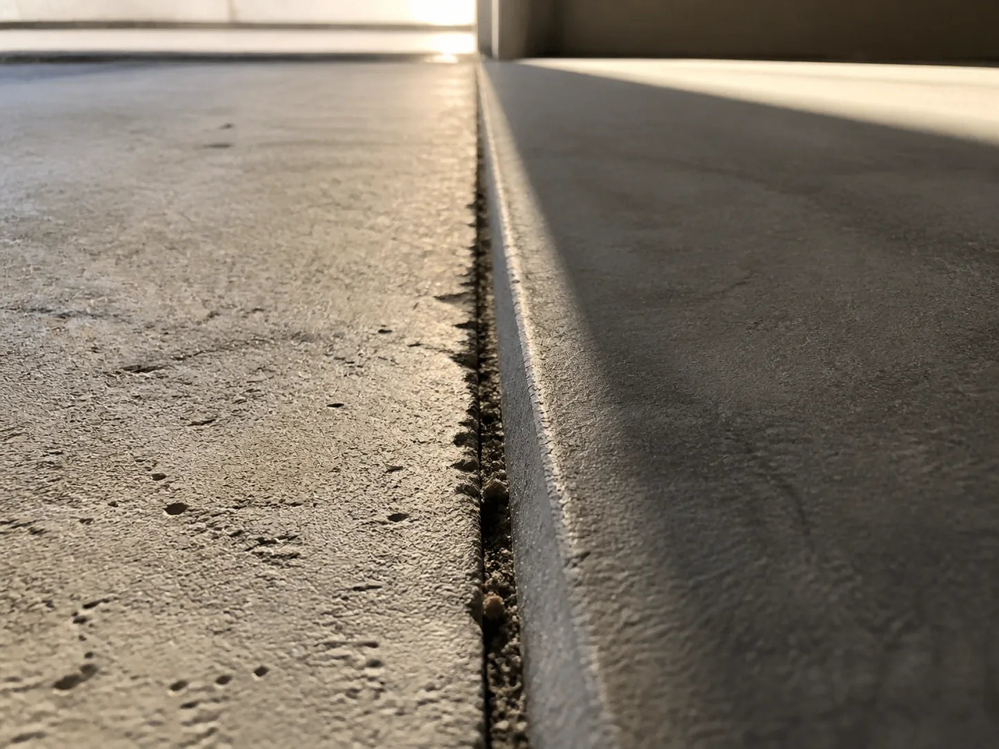 A close low angle macro of a separated slab joint at the floor transition between an original room and an added room, showing a clear vertical offset and a hairline gap
