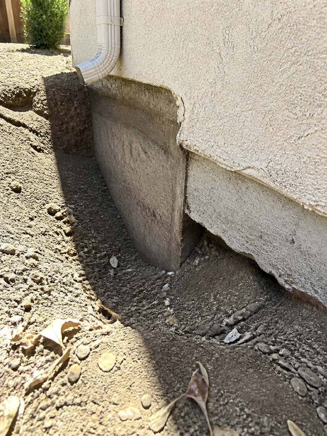 The concrete edge of a shallow addition footing exposed at a side yard where the soil grade has washed away, sitting only a few inches into the ground beside the deeper original foundation, flat harsh midday daylight