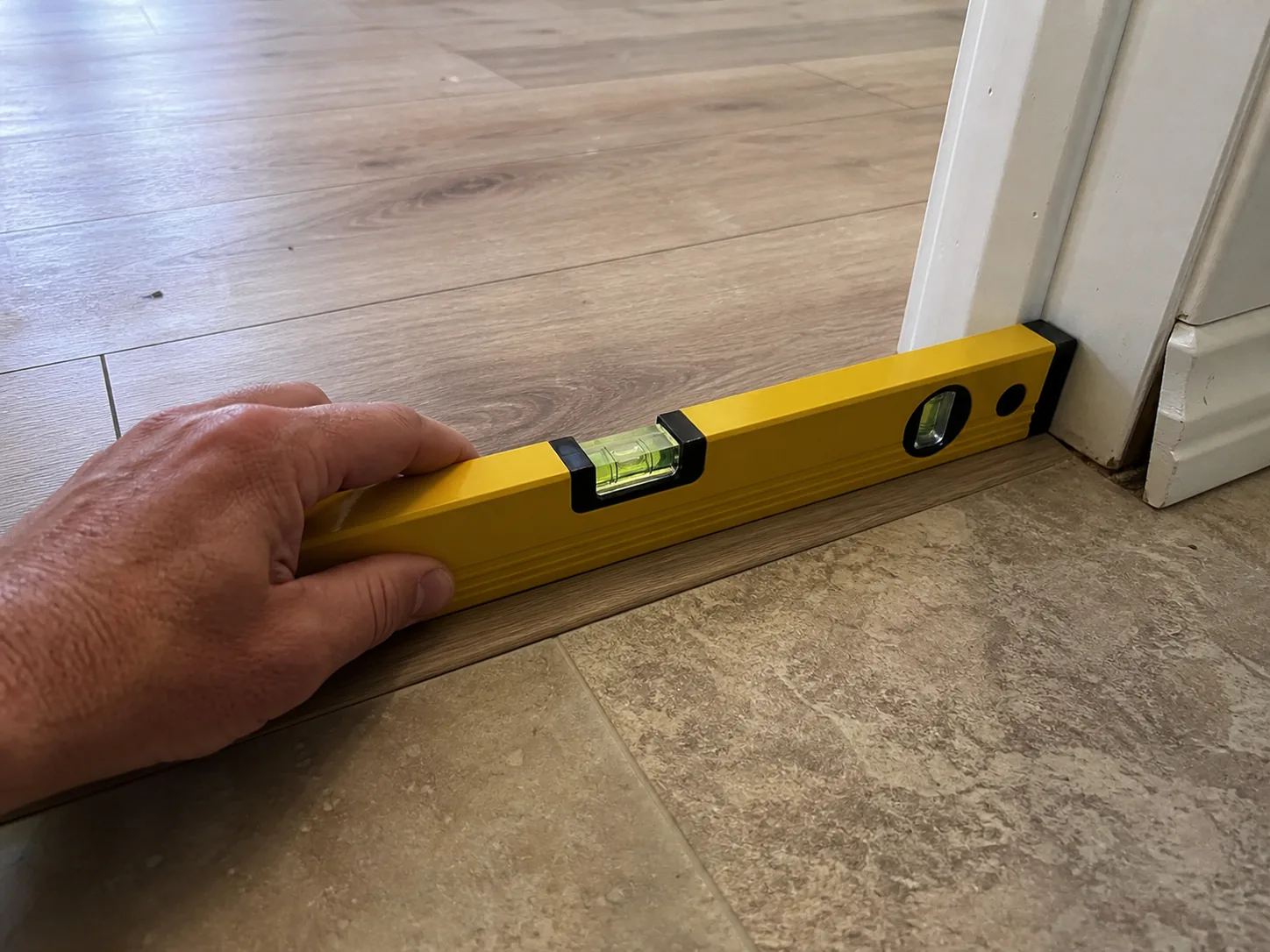 A hand seating a long spirit level across the floor threshold between an original room and an added room to check for a change in floor elevation, in available window light