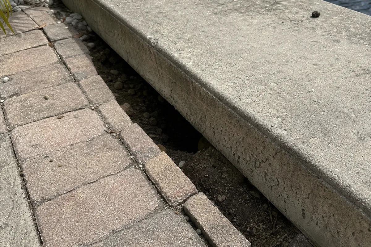 Bayfront paver walkway settled and separated just behind a concrete bulkhead cap, showing a soil void
