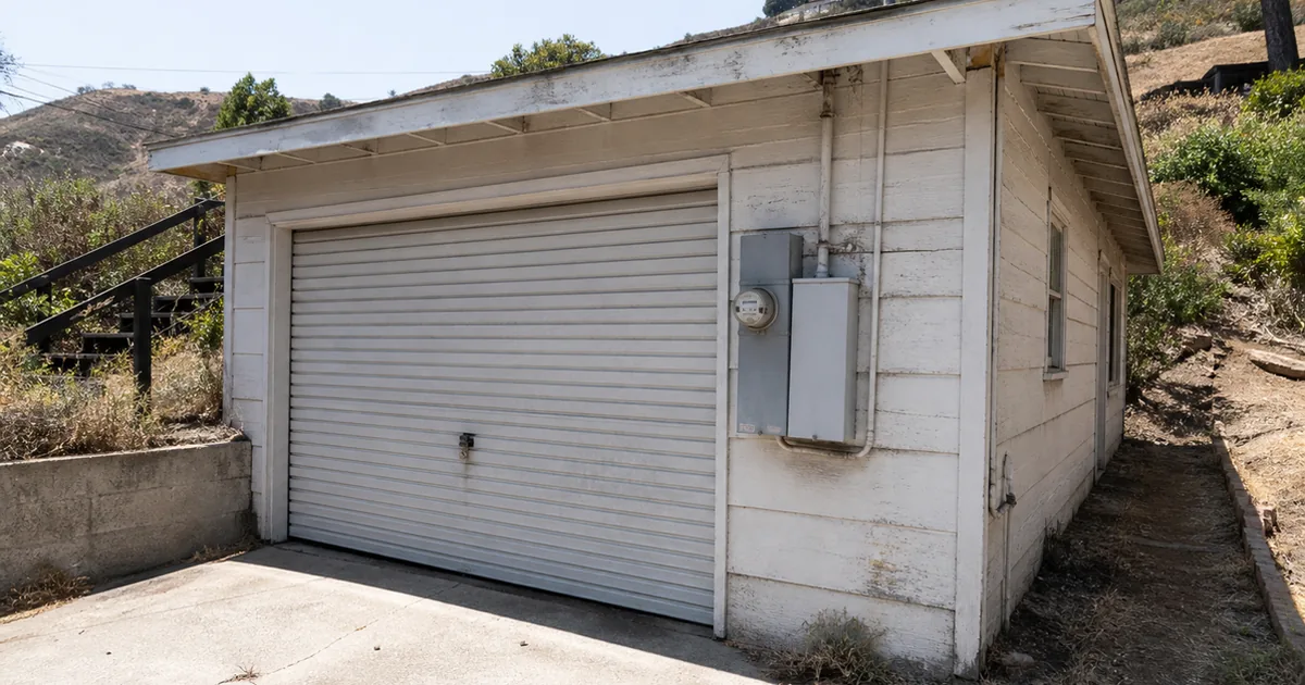 The exterior of a locked, older detached garage on a Signal Hill hillside lot, with the home's electrical meter mounted on the garage wall beside the closed door