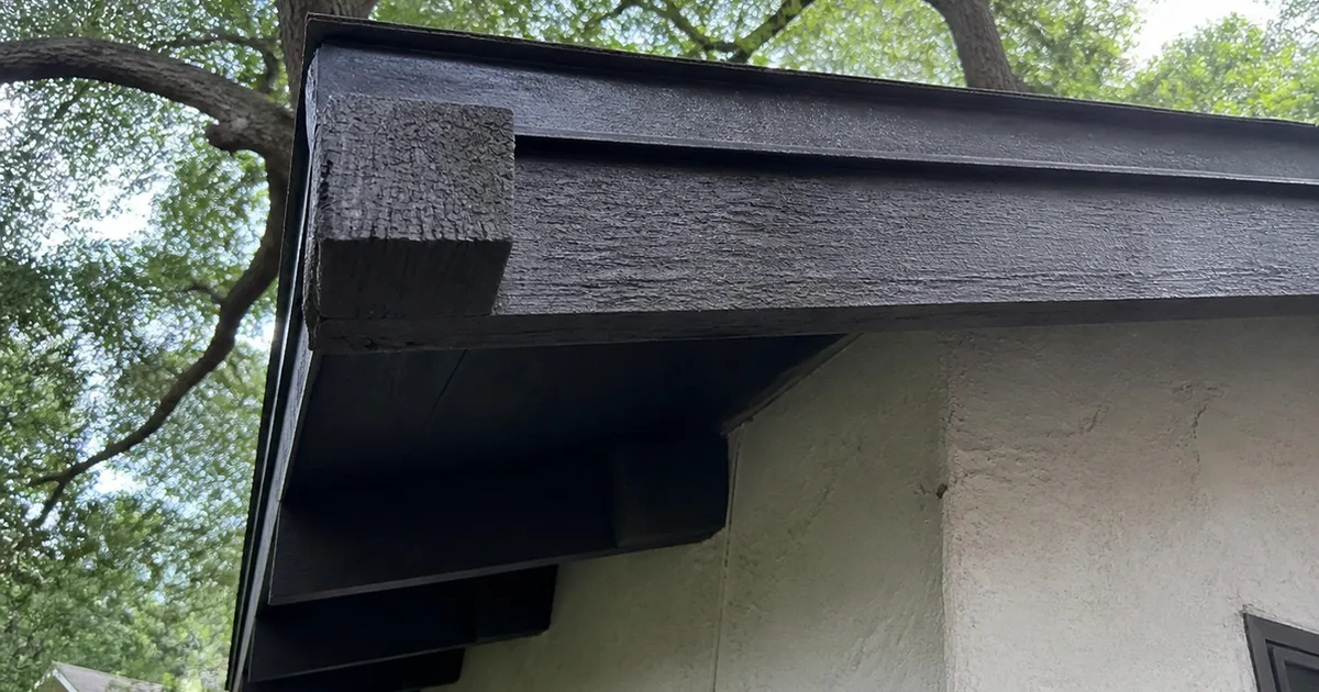 A shaded rafter tail and fascia board under a mature tree canopy on a Toluca Lake period-revival home, the wood surface dark and cracked in a cuboidal pattern