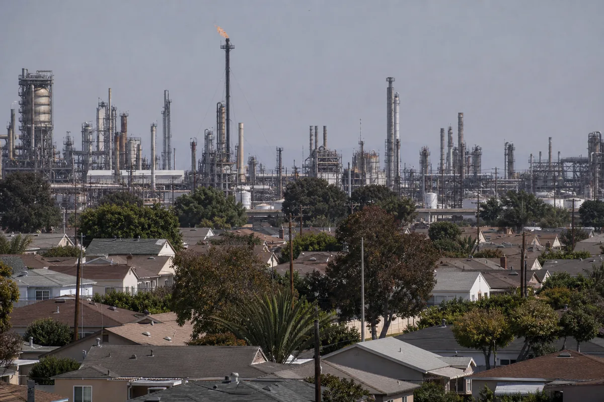 View across the rooftops of a flat South Bay residential neighborhood toward the towers, pipe racks, and flare stack of an oil refinery on the near horizon
