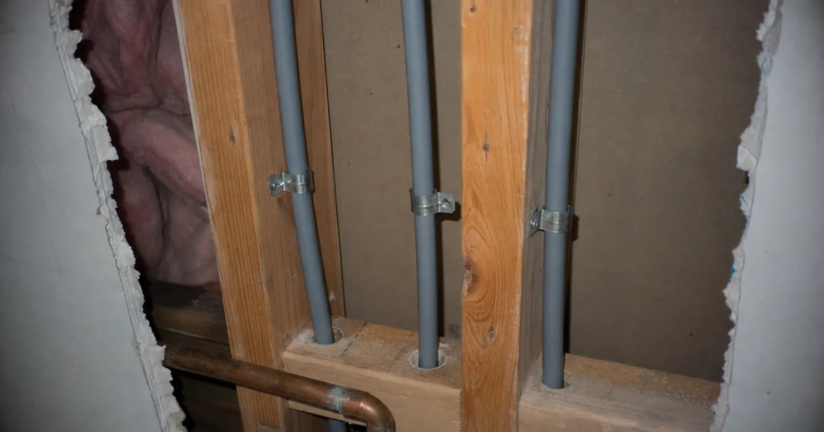 Gray polybutylene supply lines with metal clamps exposed in an opened wall cavity of a 1980s Vista tract home next to copper piping