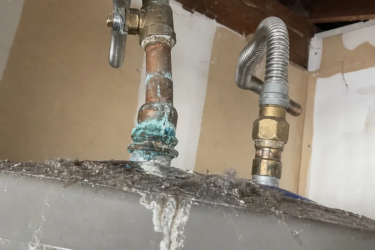 Blue-green corrosion blooming on a brass fitting at the copper supply lines above a water heater, chalky mineral trail running down