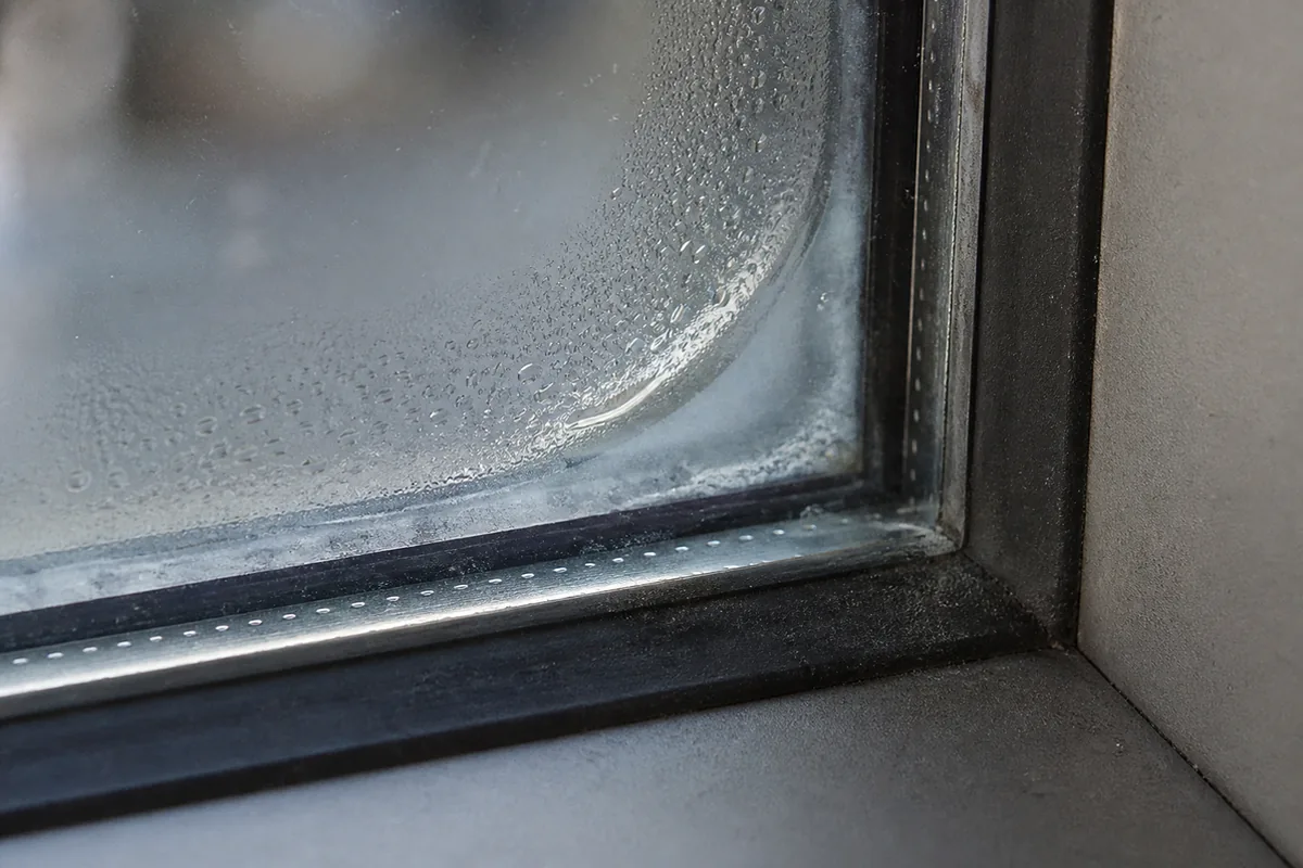 Extreme macro of the corner of a dual-pane window where the seal has failed, cloudy haze and tiny condensation droplets trapped against the metal spacer bar