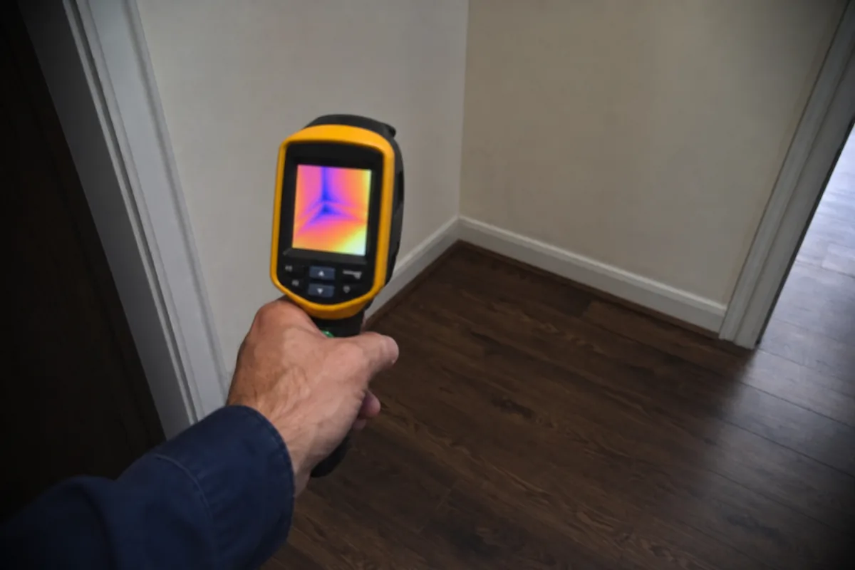 Home inspector's hand holding a handheld thermal imaging camera up to a baseboard corner to scan for heat signatures consistent with a hot-water slab leak